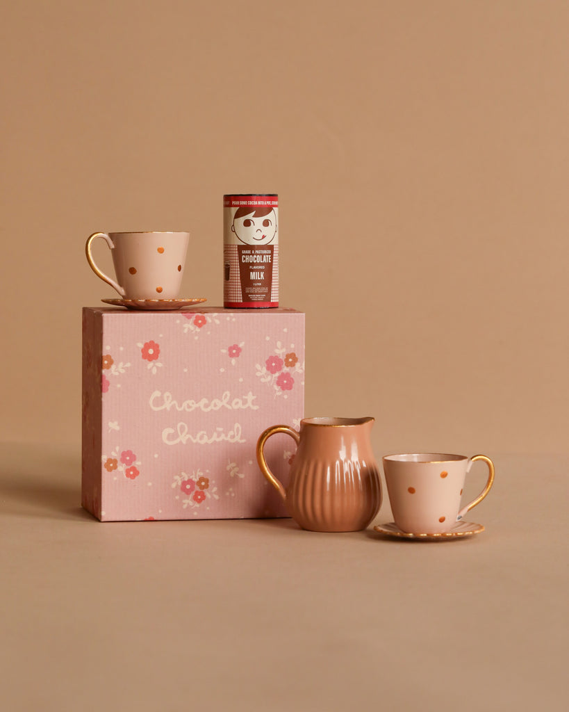 A still life featuring the Maileg Hot Chocolate Set, Mini: a pink floral "Chocolat Chaud" box with a chocolate milk powder can on top, flanked by two polka-dotted teacups from the charming Teddy family and a ribbed ceramic pitcher, all set against a warm beige background.