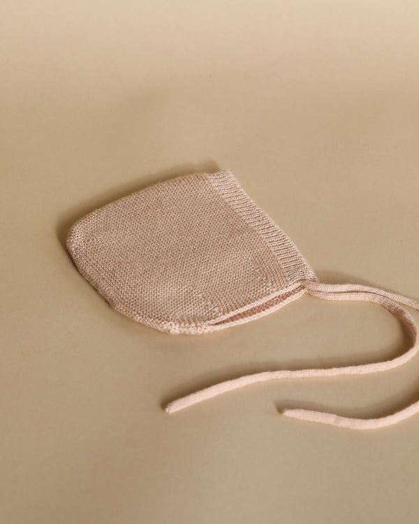 A pale pink fabric Handmade Merino Wool Newborn Bonnet - Apricot, ideal for sensitive skin, with long ties lies on a light beige background, showcasing its fine mesh texture and neatly stitched edges.