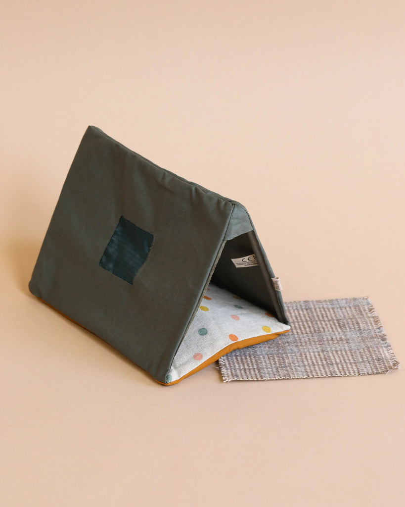 A small, tent-shaped pet bed with a dark green exterior, square window, and polka dot interior sits beside the Maileg LouiMax Little Explorer with Book Set and a woven mat on a light beige background.