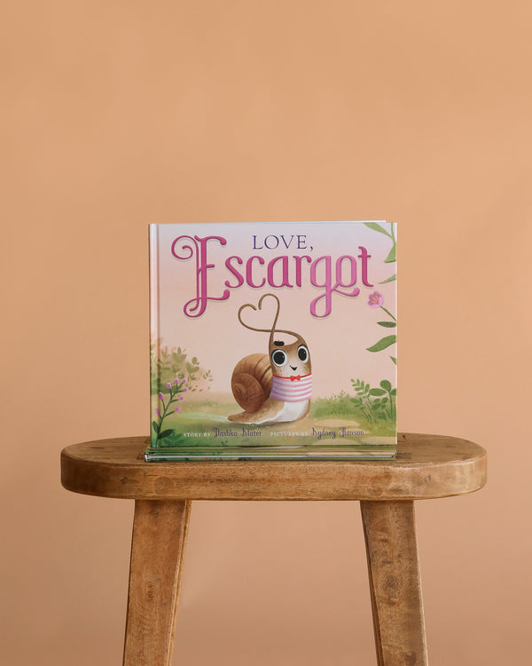 Book: Love, Escargot" is a children’s picture book from the Escargot series, featuring an illustrated snail with a heart-shaped antenna on a stool against a plain beige background.