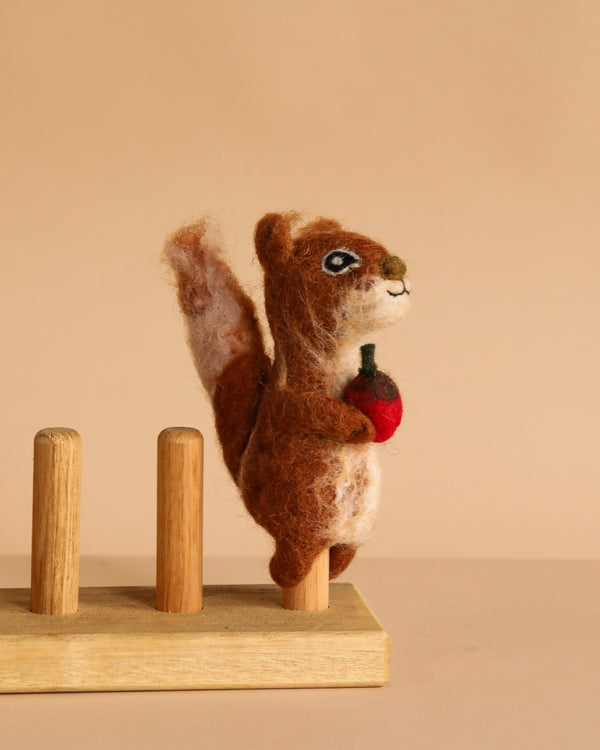 The Squirrel Finger Puppet, handcrafted by artisans in Nepal, features a brown felted squirrel standing upright and holding a small red object on a wooden base with three pegs, shown against a plain beige background.