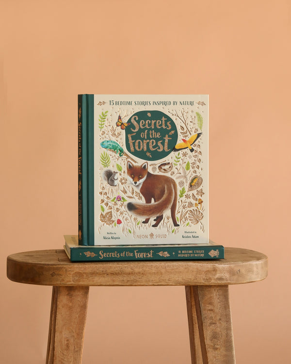 The book "Secrets Of The Forest: 15 Bedtime Stories Inspired By Nature" stands upright on a rustic wooden stool with another copy beneath it, set against a plain peach background. The illustrated cover shows woodland animals and foliage.