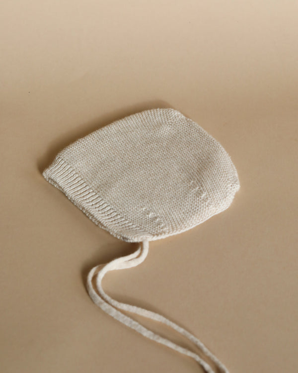 A small white Handmade Merino Wool Newborn Bonnet - Cream with a string, displayed on a beige background.