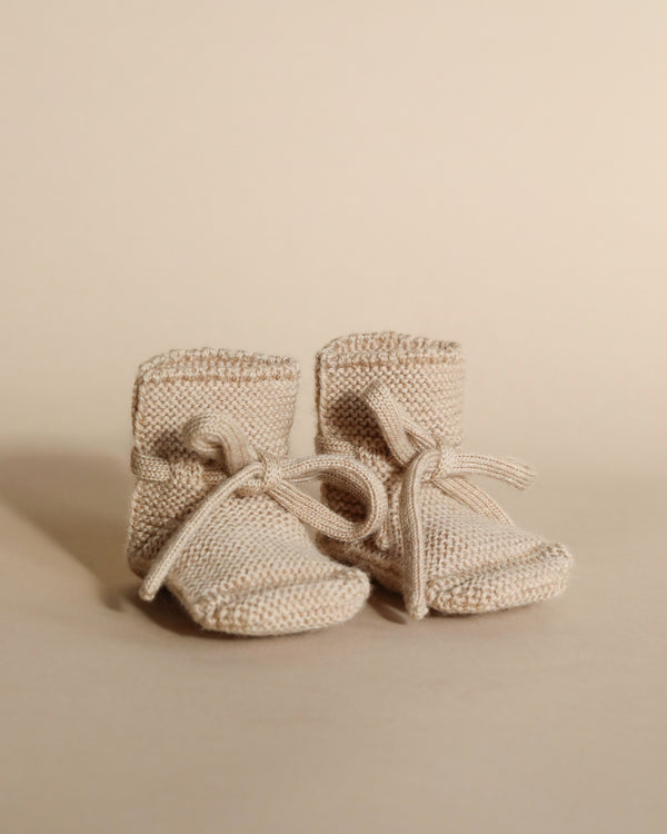 A pair of tiny, Handmade Merino Wool Booties - Sand with laces, displayed against a soft beige background. The neutral color scheme gives a warm, gentle feel to the image.