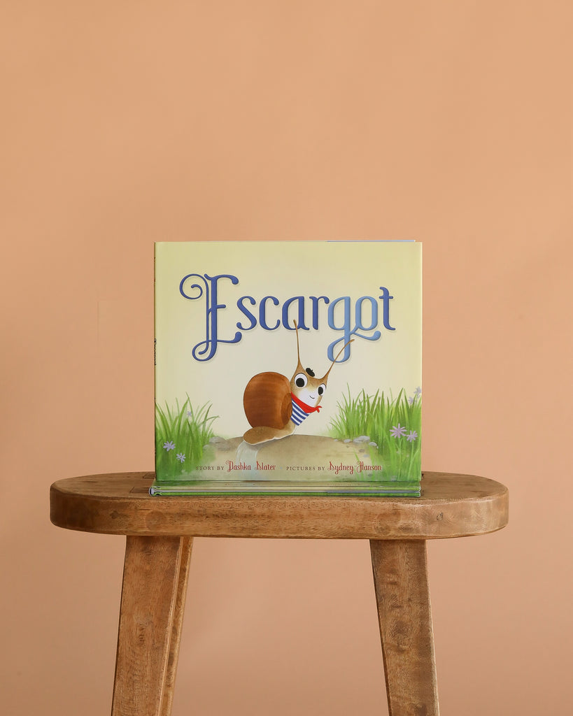 The children's picture book "Escargot" is displayed upright on a wooden stool, featuring its cover with a French snail in a striped shirt and scarf, set against a plain peach-colored background.