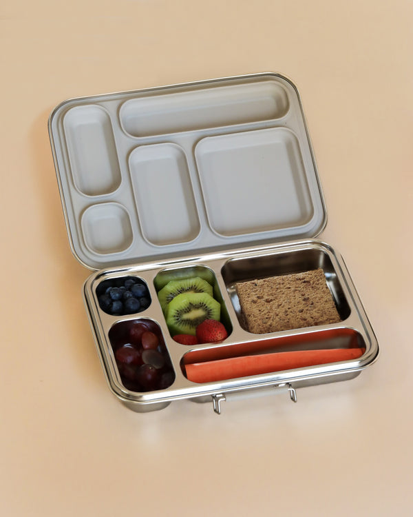 A Haps Nordic - Stainless Steel Lunch Box - Multi-Compartment (ships in approximately two weeks) open to reveal several compartments containing various meal prep foods. These include blueberries, kiwi slices, grapes, a whole strawberry, a waffle square, and a whole carrot. The background is light beige.