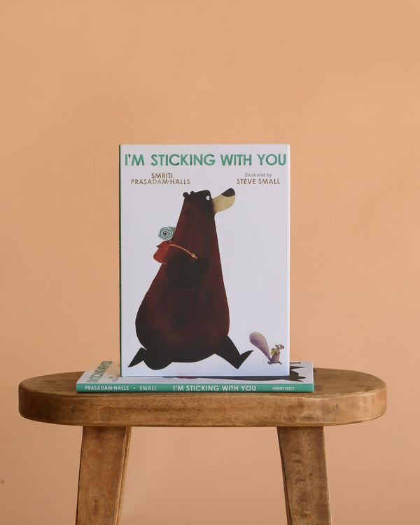 The children’s picture book "I’m Sticking With You," celebrating friendship, is shown upright on a wooden stool with another copy lying flat below, both with a bear and bird illustration, against a light orange background.