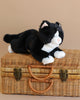 The Teddy Hermann Black Cat Stuffed Animal, featuring a white face, paws, and belly with a black body, is resting on a closed wicker suitcase. The cuddly toy slouches comfortably atop the suitcase, which has two metal latches and a woven handle. The scene is set against a neutral beige background.