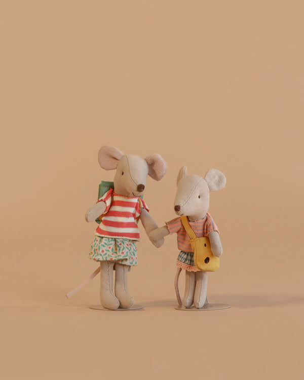 Two Maileg Sister Mice stand side by side against a beige background. One wears a striped shirt, skirt, and backpack; the other has on plaid and carries a yellow satchel. The Maileg Sister Mice Set appears to be holding hands.