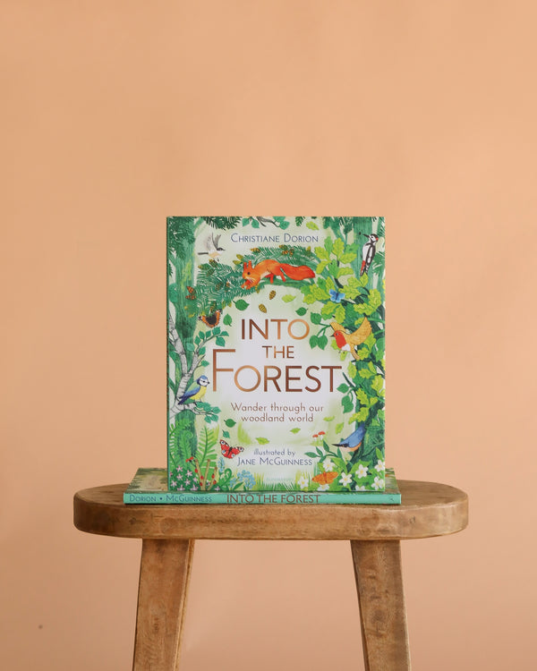 The book "Into The Forest" stands upright on a wooden stool against a peach background, its colorful cover featuring woodland animals and lush foliage, inviting readers to discover the wonders of the forest.