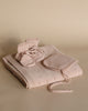 A neatly arranged set of baby clothes including a Handmade Merino Wool Bibi Blanket - Apricot, a pair of booties, and a hat, all in a soft pink color on a beige background.