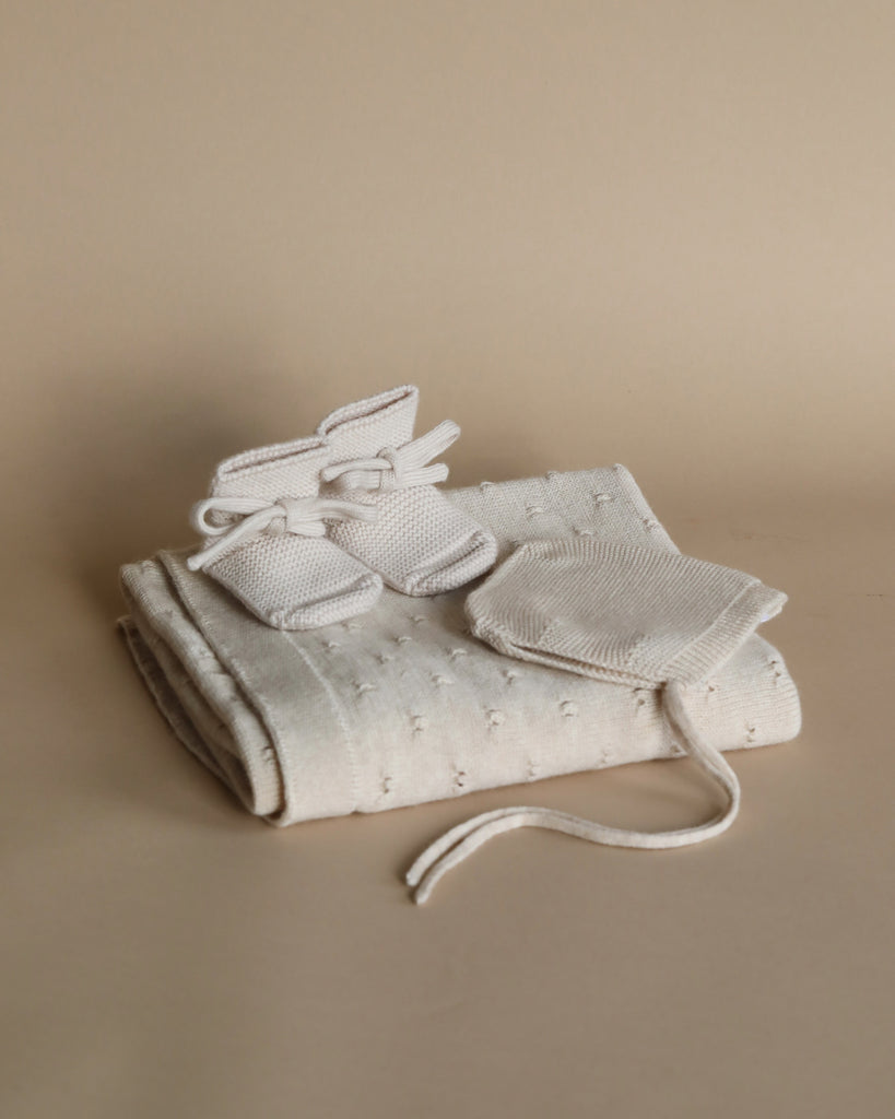 A pair of small white knit baby booties placed atop a folded Handmade Merino Wool Bibi Blanket - Cream, all set against a neutral beige background.