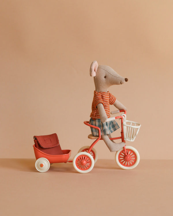 Maileg Big Sister’s Cycling Adventure features a fabric mouse in a striped shirt and plaid shorts riding a red tricycle with white handles, pulling a red wagon and brown bag against a beige background—a perfect Mouse Accessories addition.