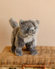 A Teddy Hermann Gray Standing Cat Stuffed Animal from the Hermann Teddy Collection stands atop a wicker basket, positioned against a plain beige backdrop. With black eyes and a slightly darker face and legs, this child-friendly design adds character to its soft appearance.