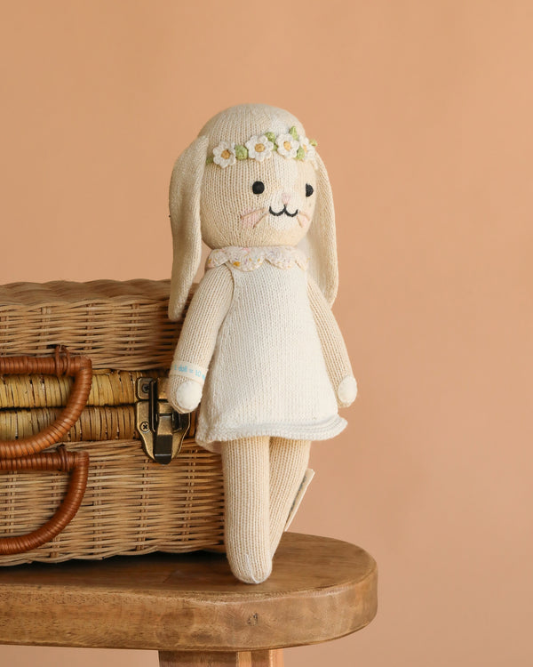 Cuddle + Kind Hannah the Bunny (Ivory Floral Crown), a hand-knit doll in a white dress and floral headband, sits on a wicker basket atop a wooden stool against a peach background, making a charming cotton stuffed animal.