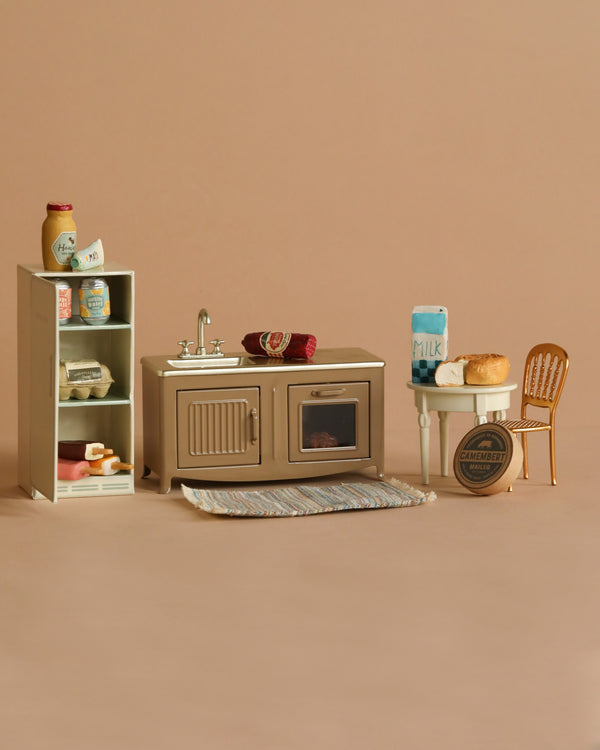 The Maileg Kitchen Set includes a fridge with jam and bread, a counter with sink and oven, and a cozy breakfast-ready table. A Maileg Striped Rug enhances the miniature scene against its warm beige backdrop.