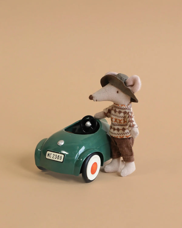 The Maileg Big Brother Mouse Holiday Adventure Set features a mouse in a sweater, brown pants, and hat beside a green car with "MC 2389" license plate and cute accessories, ready for adventure on a beige background.