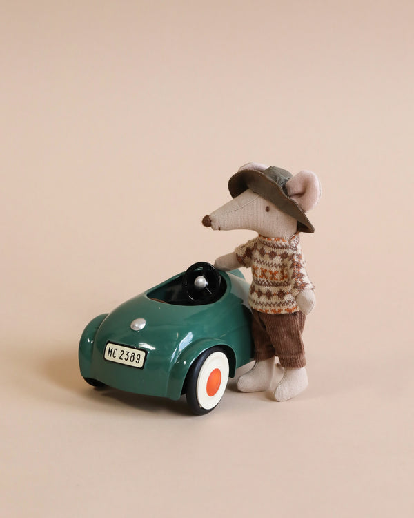 A Maileg Mouse Car - Dark Green, dressed in a knitted sweater and a hat, sitting in against a beige background.
