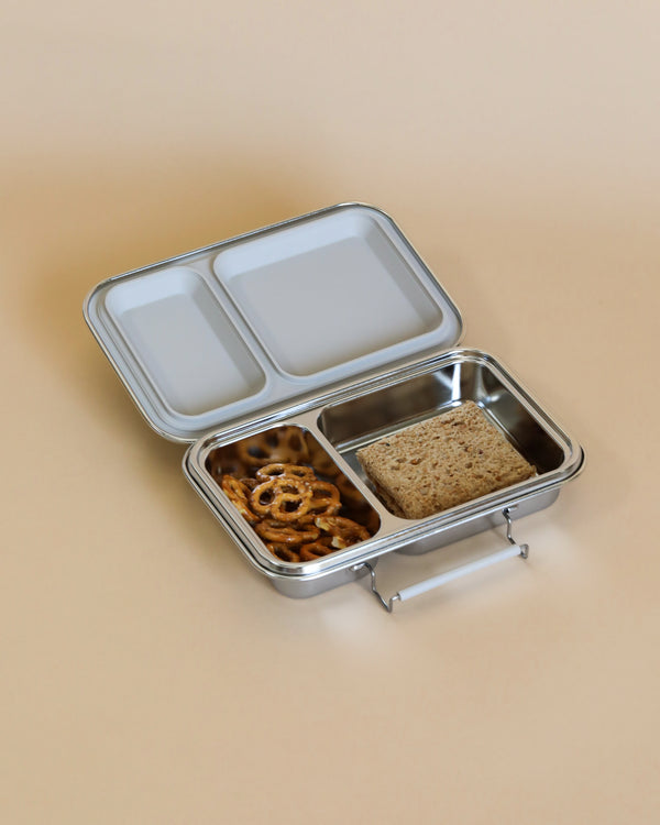 A stainless steel Haps Nordic - Stainless Steel Lunch - Two Compartments lunchbox with separated compartments: pretzels in one, a square sandwich in another, and an empty top compartment. The lunchbox is on a beige background, with one lid open to reveal the food inside.