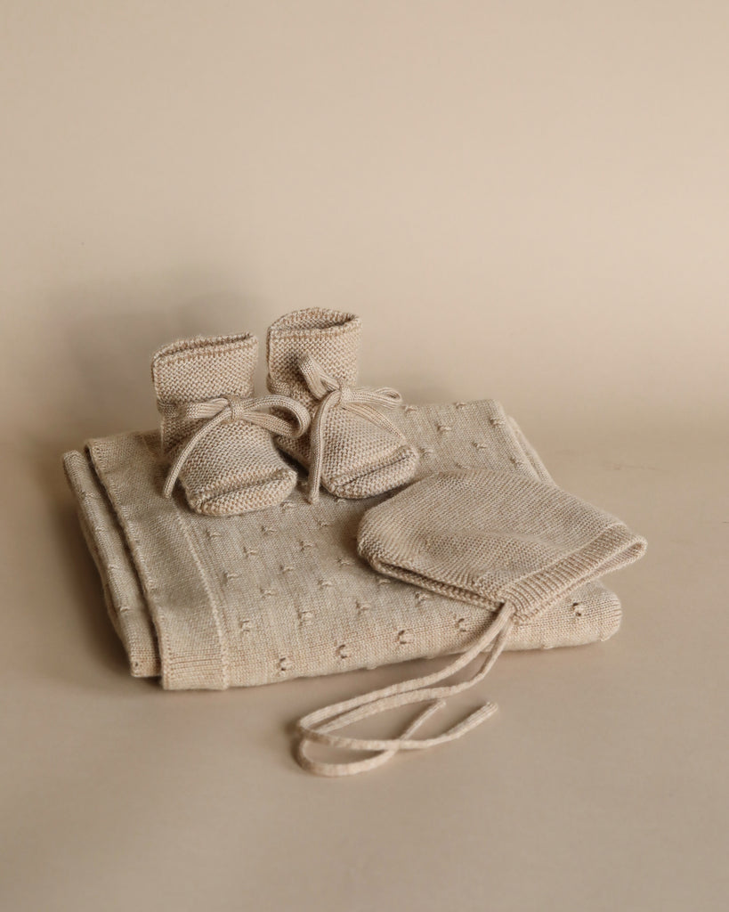 A pair of small, eczema-friendly Handmade Merino Wool Booties - Sand on top of a matching blanket, all in a soft beige color, against a plain, light beige background.