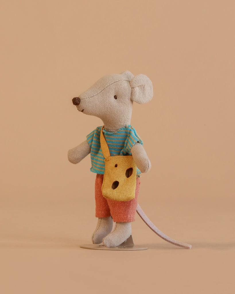 A Maileg Little Brother Adventure Set mouse, dressed in a blue striped shirt, orange shorts, and carrying a yellow cheese-shaped bag, stands on a neutral beige background. This charming Adventure Set mouse is perfect for imaginative play and storytelling.