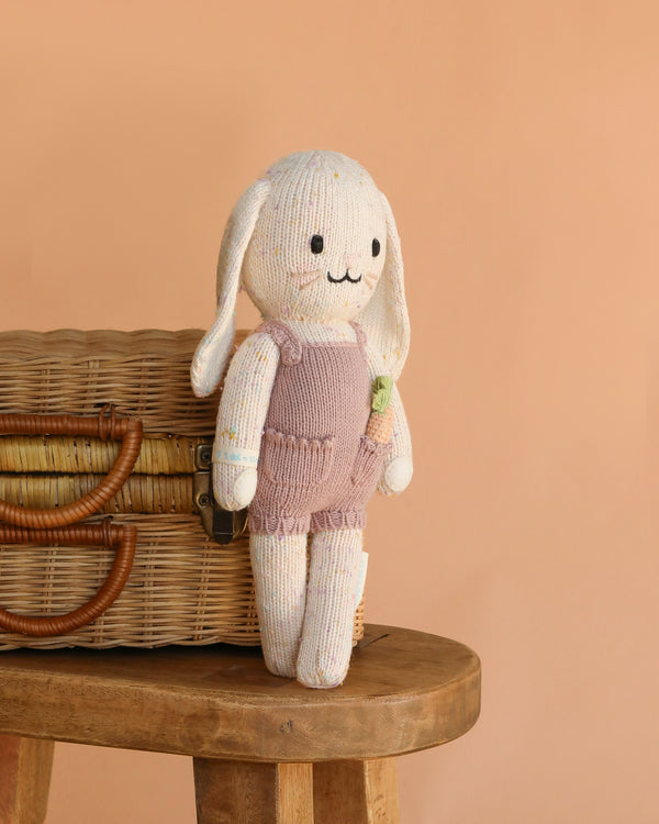 Cuddle + Kind Harper the Bunny, a hand-knit doll with floppy ears and a pink romper made from premium cotton yarn, stands on a wooden stool beside a wicker basket against a peach backdrop.
