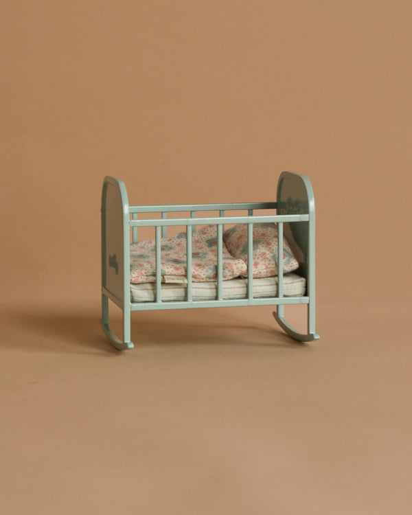 The Maileg My Cradle - Blue Mint, featuring ornate metal side panels and floral-patterned bedding, is set against a plain beige background. This small, vintage-style cradle is empty, evoking a nostalgic or decorative theme.