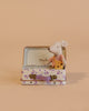 The Maileg Little Sister On the Go Suitcase Set features a mouse in a pink outfit holding cheese, sitting inside an open floral suitcase with a lavender handle. Not suitable for children under three due to small parts.