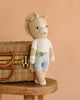 Cuddle + Kind Liam the Pony, a fair trade, hand-knit plush doll in light blue shorts and a white shirt, leans against a wicker basket on a wooden stool with a plain beige background.