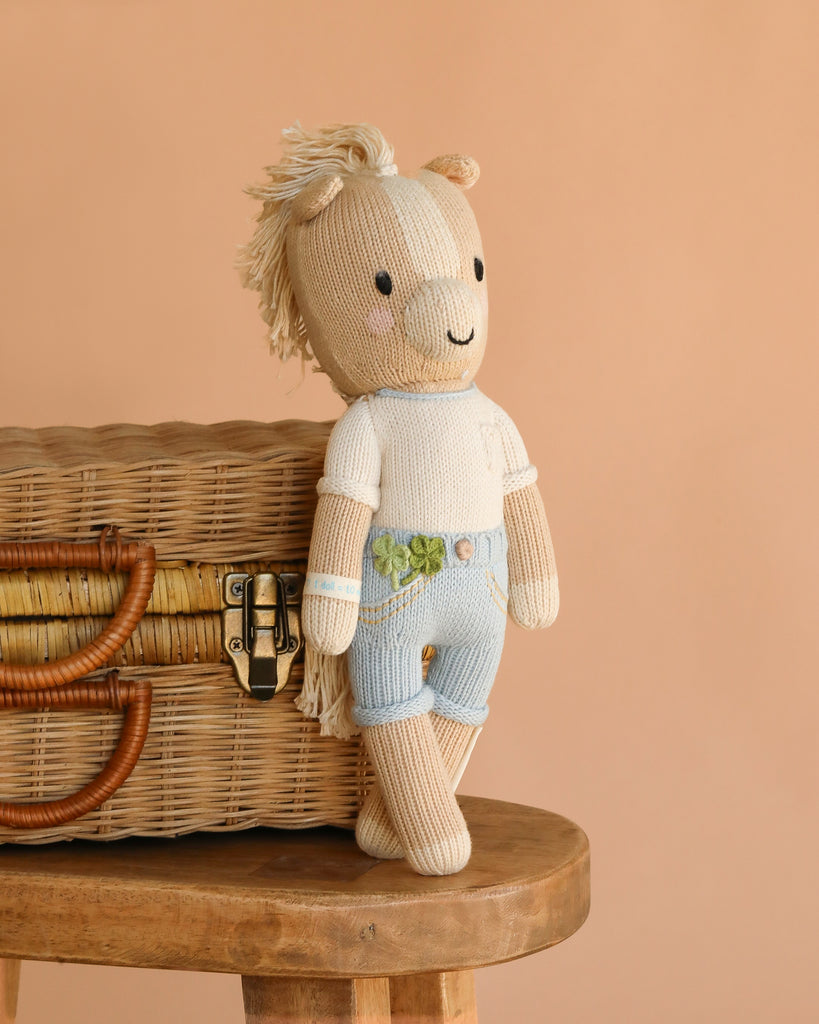 Cuddle + Kind Liam the Pony, a fair trade, hand-knit plush doll in light blue shorts and a white shirt, leans against a wicker basket on a wooden stool with a plain beige background.