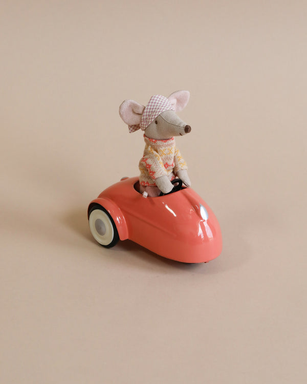 A Maileg Mouse Car - Coral figurine of a mouse wearing a plaid hat and sweater, driving a shiny red metal car against a plain beige background.