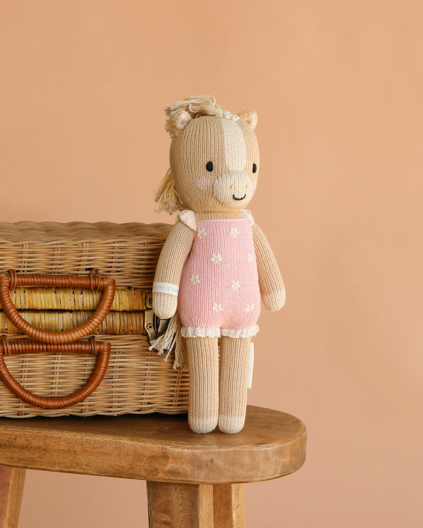 The Cuddle + Kind Sophia the Pony, a handmade doll with a beige body and pink outfit with white stars, leans against a woven basket on a wooden stool against a peach background. Ethically handcrafted in Peru.