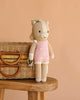 The Cuddle + Kind Sophia the Pony, a handmade doll with a beige body and pink outfit with white stars, leans against a woven basket on a wooden stool against a peach background. Ethically handcrafted in Peru.