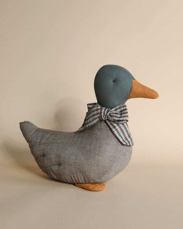 A handmade Maileg Duck Stuffed Animal - Gray with a blue head, orange beak, and grey body, adorned with a plaid bow tie suitable for festive season décor, set against a plain beige background.