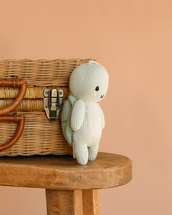 The Cuddle + Kind Baby Sea Turtle (Kelp Green), a small hand-knit plush toy, sits on a wooden stool beside a closed wicker basket with leather straps, set against a plain peach background.