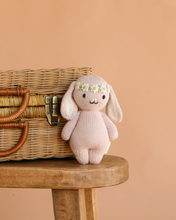 The Cuddle + Kind Baby Bunny, Rose (Ivory Floral Crown), a hand-knit plush with long ears and a floral crown made from soft Peruvian cotton yarn, sits on a wooden stool beside a wicker basket against a peach background.