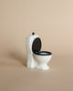 A small, white Maileg LouiMax Bathroom Set toilet with an open black lid sits on a beige surface against a plain beige background, perfectly sized for your Maileg mouse or to pair with a Maileg sink dresser.