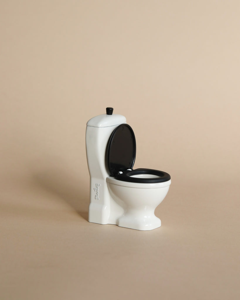 A small, white Maileg LouiMax Bathroom Set toilet with an open black lid sits on a beige surface against a plain beige background, perfectly sized for your Maileg mouse or to pair with a Maileg sink dresser.