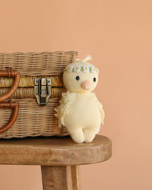 The Cuddle + Kind Baby Duckling (Ivory Floral Crown) is an heirloom-quality, hand-knit plush made from soft Peruvian cotton yarn, shown perched on a wooden stool by a wicker basket against a peach background.