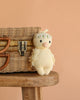 The Cuddle + Kind Baby Duckling (Ivory Floral Crown) is an heirloom-quality, hand-knit plush made from soft Peruvian cotton yarn, shown perched on a wooden stool by a wicker basket against a peach background.