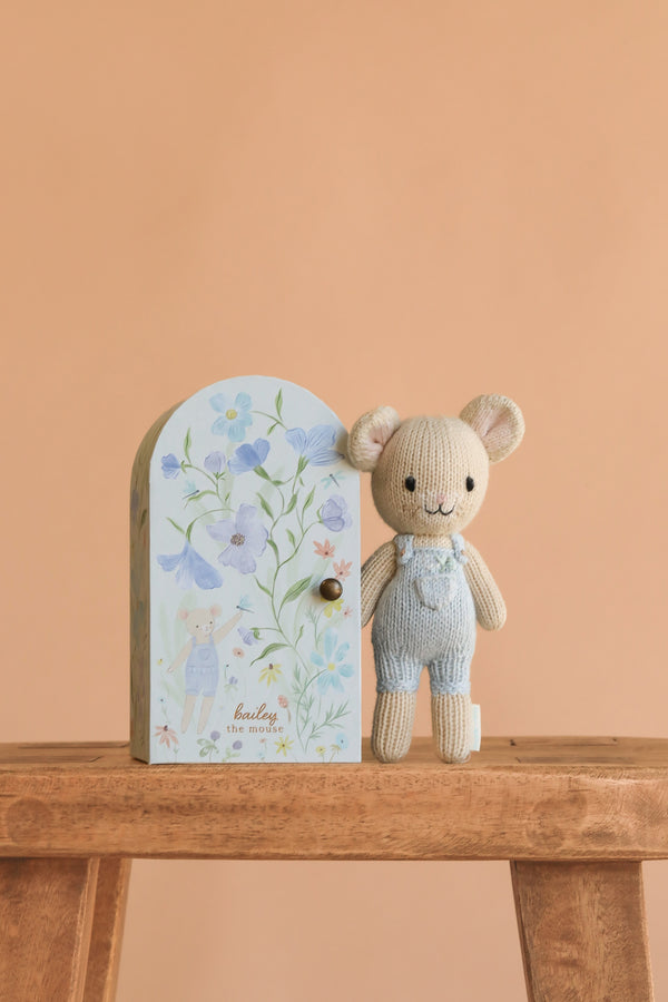 Cuddle + Kind’s Tiny Bailey the Mouse, hand knit in Peru and dressed in blue overalls, sits by a floral pastel box with matching art on a wooden stool against a peach background—a charming, sustainable, fair trade gift.