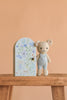 Cuddle + Kind’s Tiny Bailey the Mouse, hand knit in Peru and dressed in blue overalls, sits by a floral pastel box with matching art on a wooden stool against a peach background—a charming, sustainable, fair trade gift.