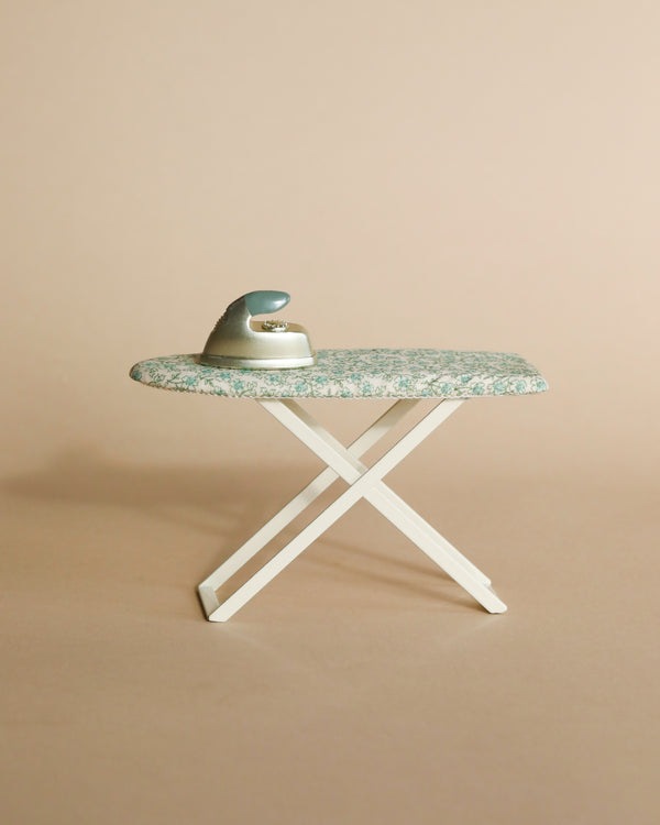 A vintage iron resting on a small Maileg Ironing - Mouse Size set against a plain beige background. The ironing board is folded open and positioned upright.