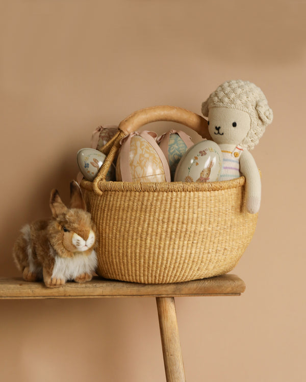 A Large Easter Egg Basket Set with decorative eggs and a Cuddle + Kind Lamb toy sits on a wooden bench, while a plush rabbit rests nearby against a beige background, showcasing fair trade practices.