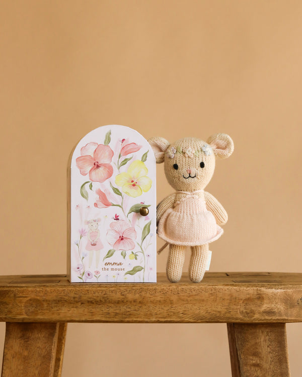 Cuddle + Kind Tiny Emma the Mouse, a hand-knit doll in a pink dress, stands on a wooden bench beside a pastel floral box featuring a mouse illustration, handmade in Peru and set against a plain beige background.