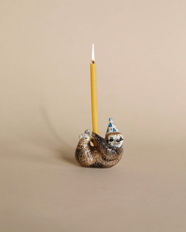 A lit yellow candle standing in a whimsical, hand-painted Sloth Cake Topper holder with a party hat against a beige background.