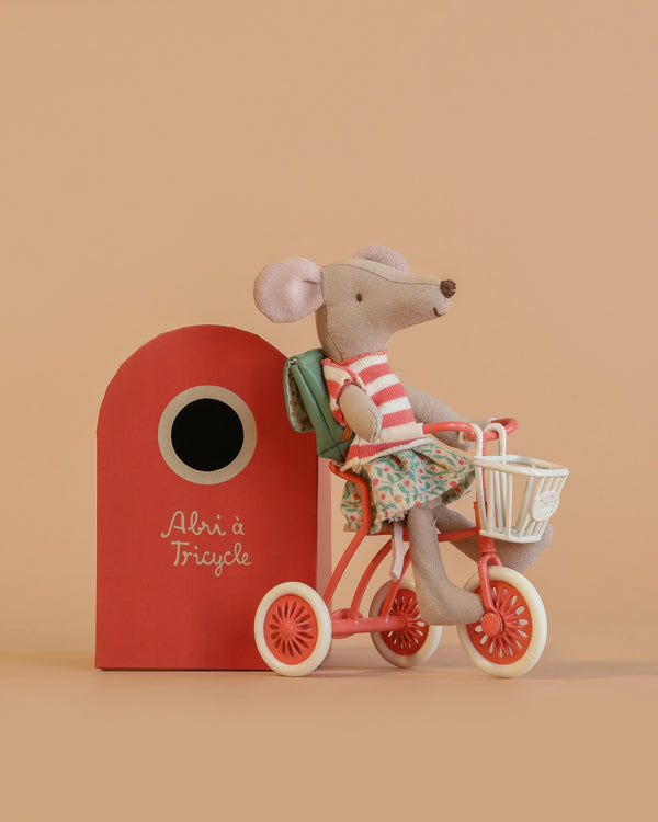 The Maileg Big Sister Tricycle Set features a mouse in a striped shirt and floral skirt riding a red tricycle next to the red "Abri à Tricycle" box, all displayed against a beige background.