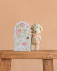 Cuddle + Kind Tiny Lucy the Lamb, a hand-knit newborn-safe doll with a pink bow, stands on a wooden bench next to an illustrated door labeled “Lucy the lamb,” set against a peach background. Perfect for gentle play.