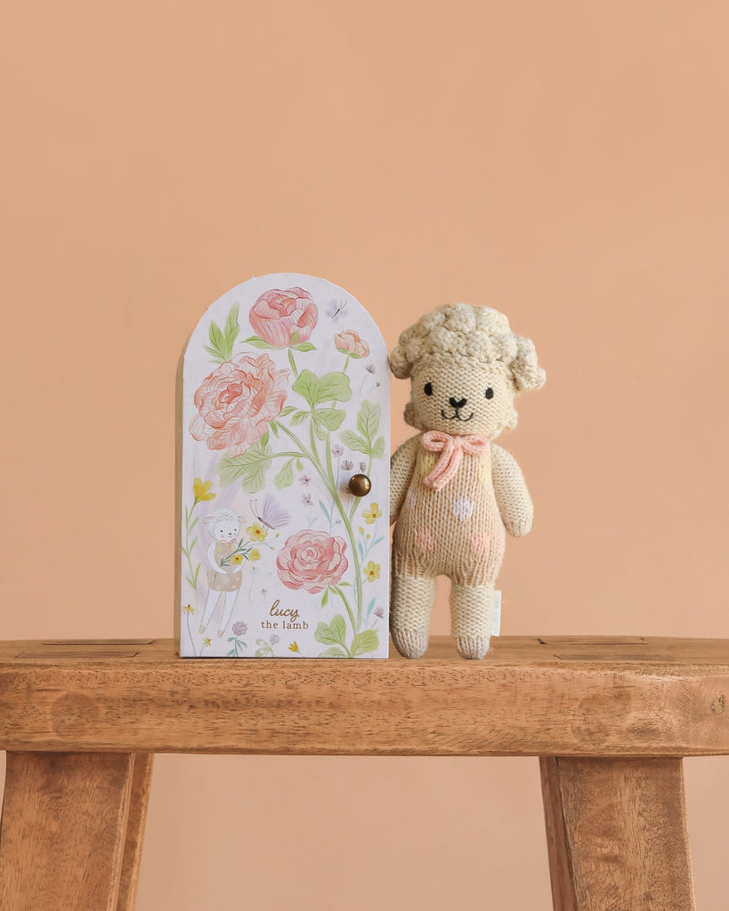 Cuddle + Kind Tiny Lucy the Lamb, a hand-knit newborn-safe doll with a pink bow, stands on a wooden bench next to an illustrated door labeled “Lucy the lamb,” set against a peach background. Perfect for gentle play.