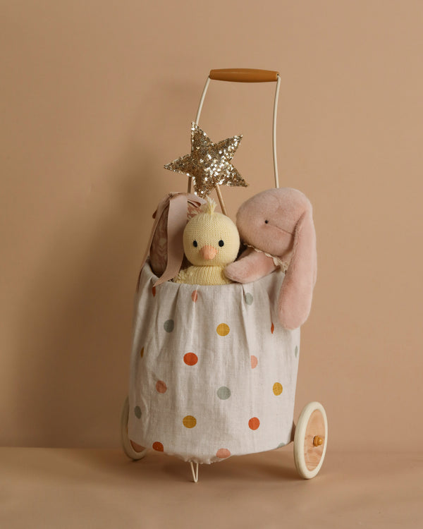 The Easter Trolley Set features a wooden-handled toy cart with a polka-dotted fabric exterior and white wheels on a beige background, holding soft toys like a yellow knit doll, plush pink bunny, and Gold Sequin Star Wand.